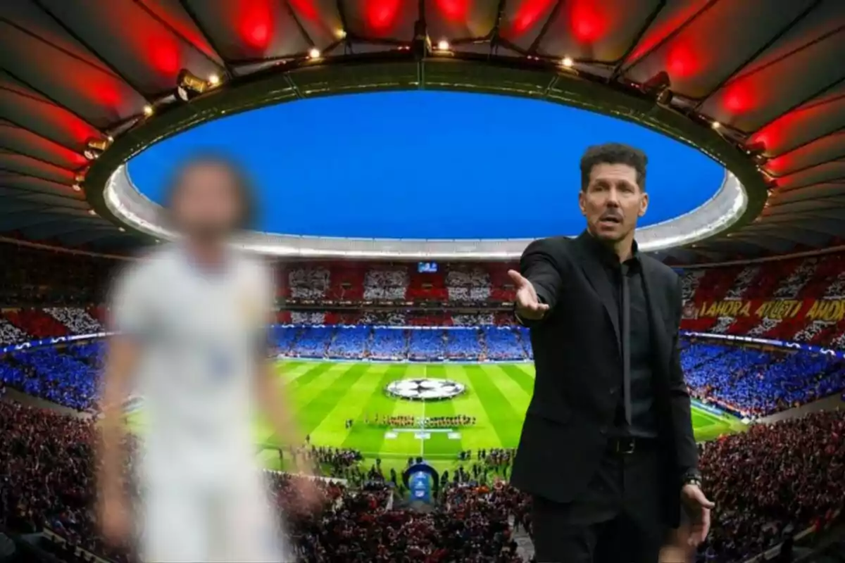 Un entrenador de fútbol gesticula en el campo mientras un jugador desenfocado se encuentra en primer plano, con un estadio lleno de aficionados de fondo.