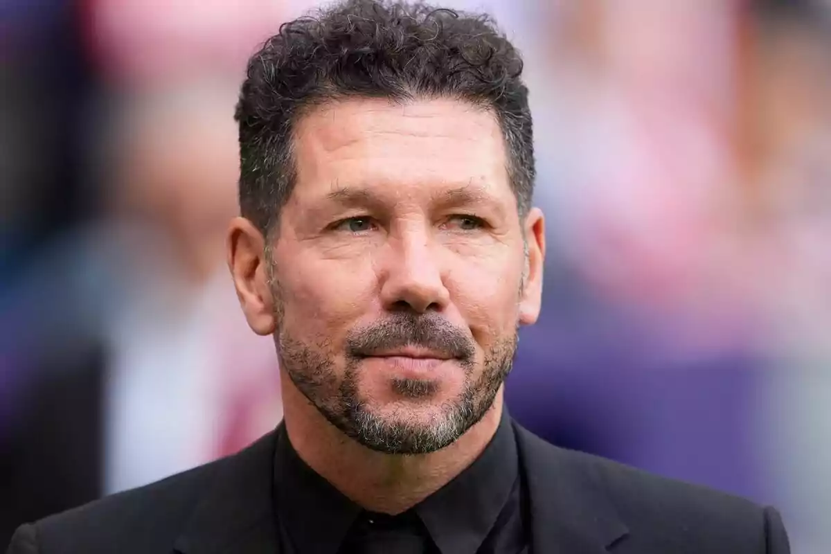 Un hombre con cabello oscuro y barba corta viste un traje negro y mira hacia adelante con una expresión seria.