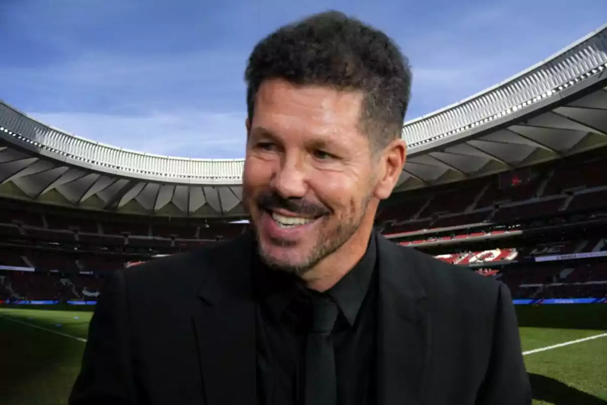 Un hombre sonriente con barba y cabello corto, vestido con traje negro, en un estadio de fútbol.