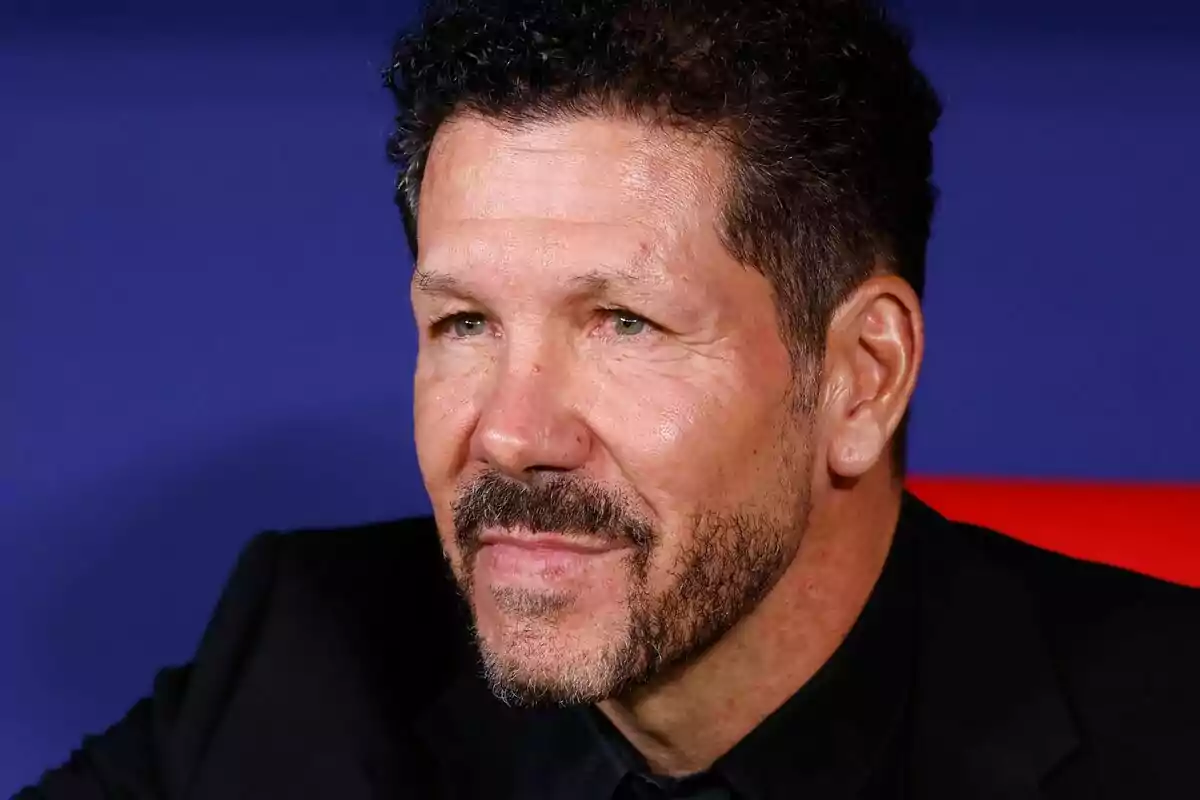 Un hombre con barba y cabello rizado mira hacia un lado con una expresión seria y un fondo azul.