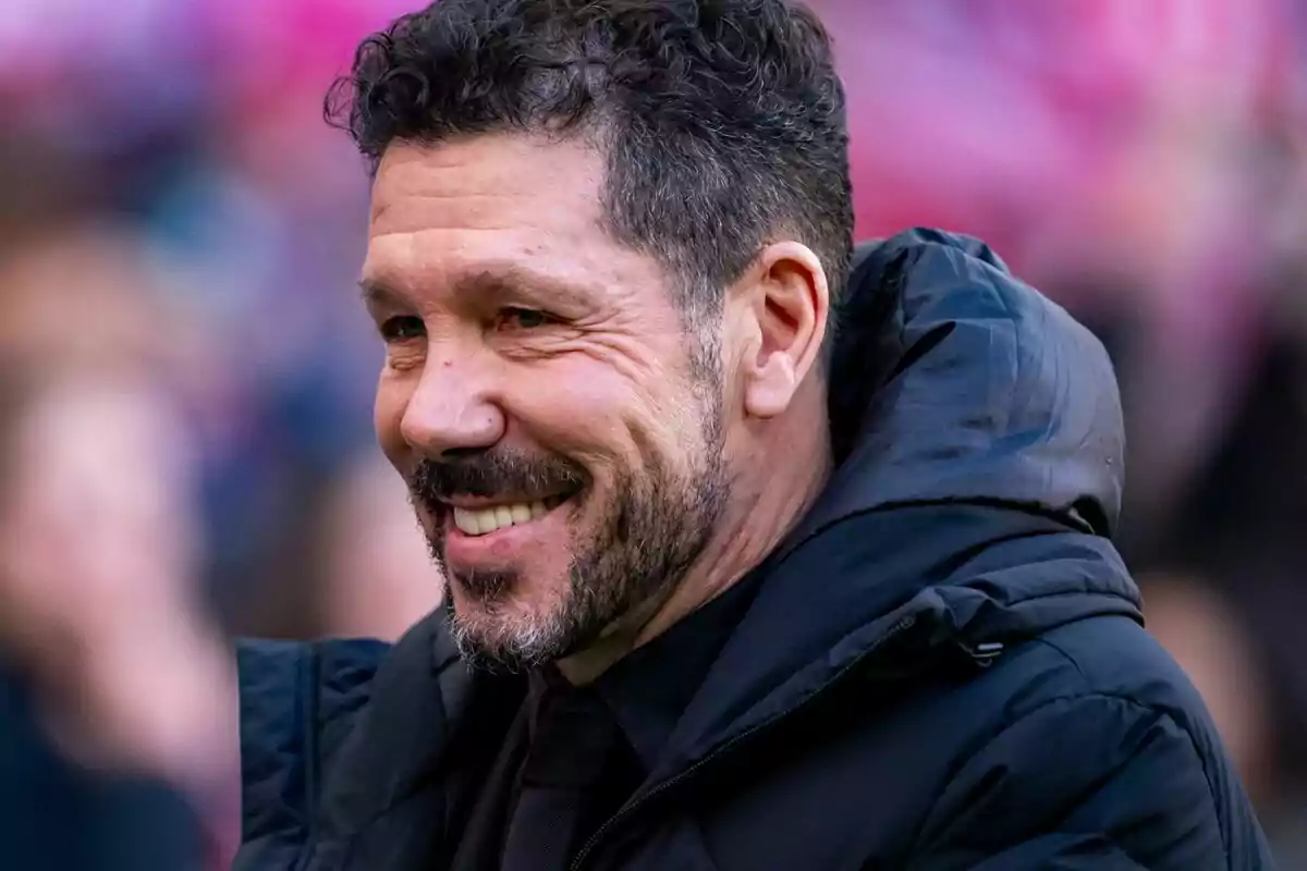 Un hombre sonriente con barba y cabello rizado lleva una chaqueta oscura.