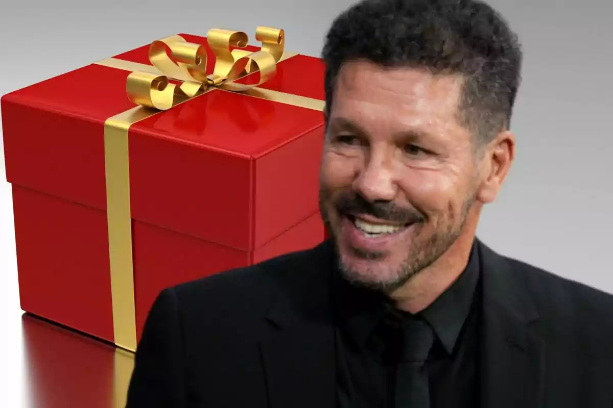 Un hombre sonriente con barba y cabello corto, vestido de traje negro, frente a una caja de regalo roja con lazo dorado.