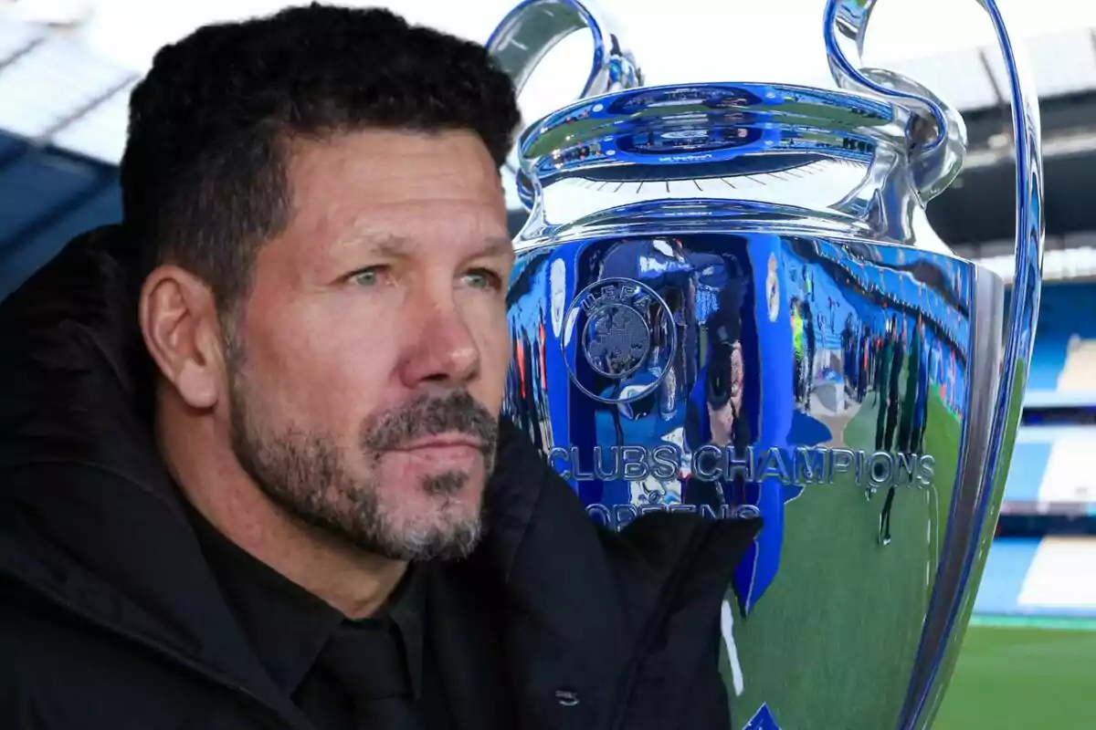 Un hombre con barba y cabello oscuro está junto a un gran trofeo plateado en un estadio de fútbol.