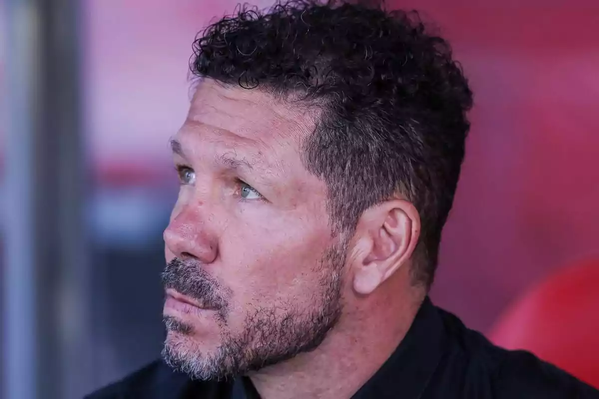 Hombre de cabello rizado y barba mirando hacia la izquierda con expresión seria y fondo desenfocado en tonos rojos