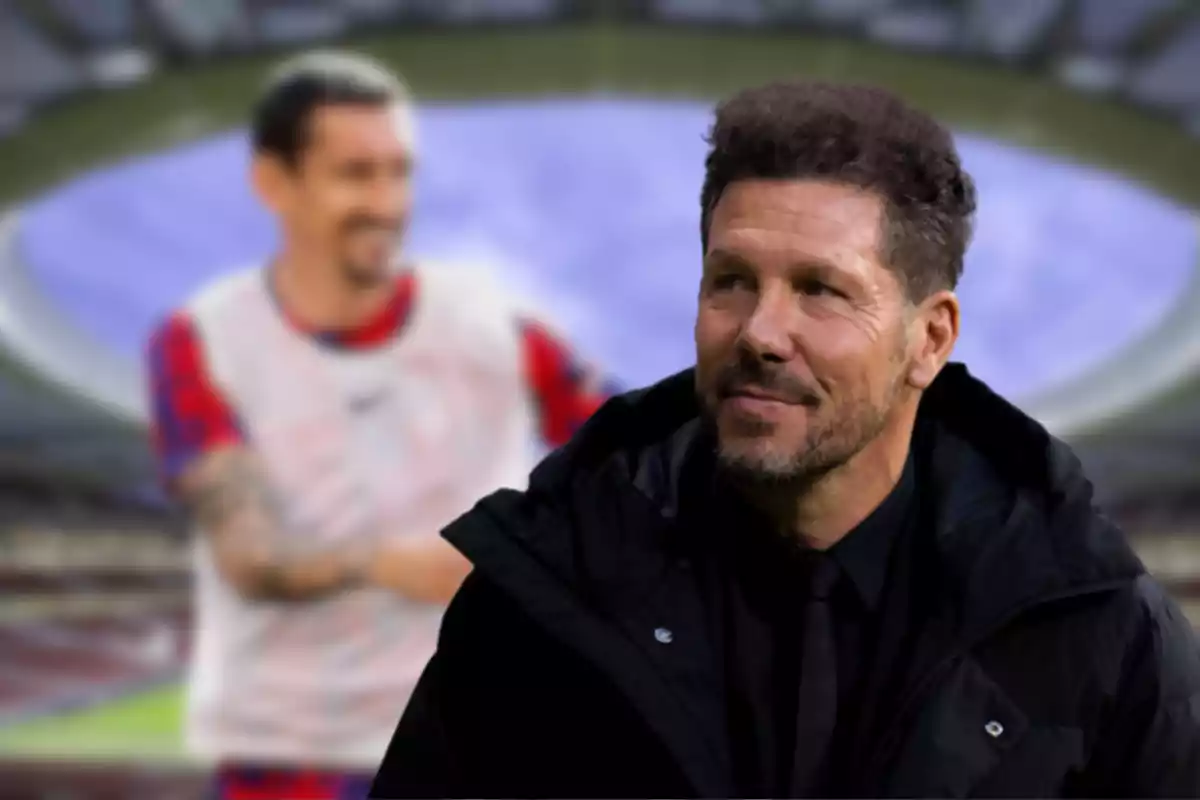 Un hombre con chaqueta negra y barba sonríe mientras está de pie en un estadio de fútbol, con una persona desenfocada en el fondo.