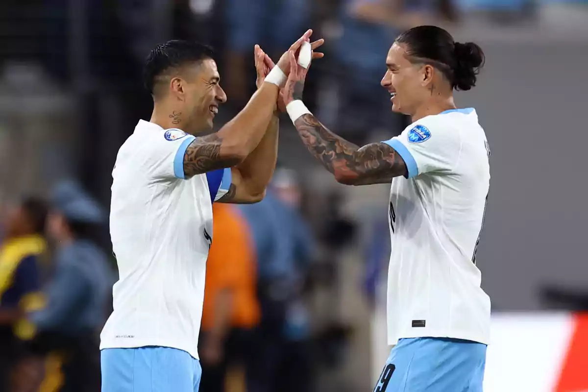 Dos futbolistas con uniformes blancos y celestes se dan un saludo de manos en el campo de juego.