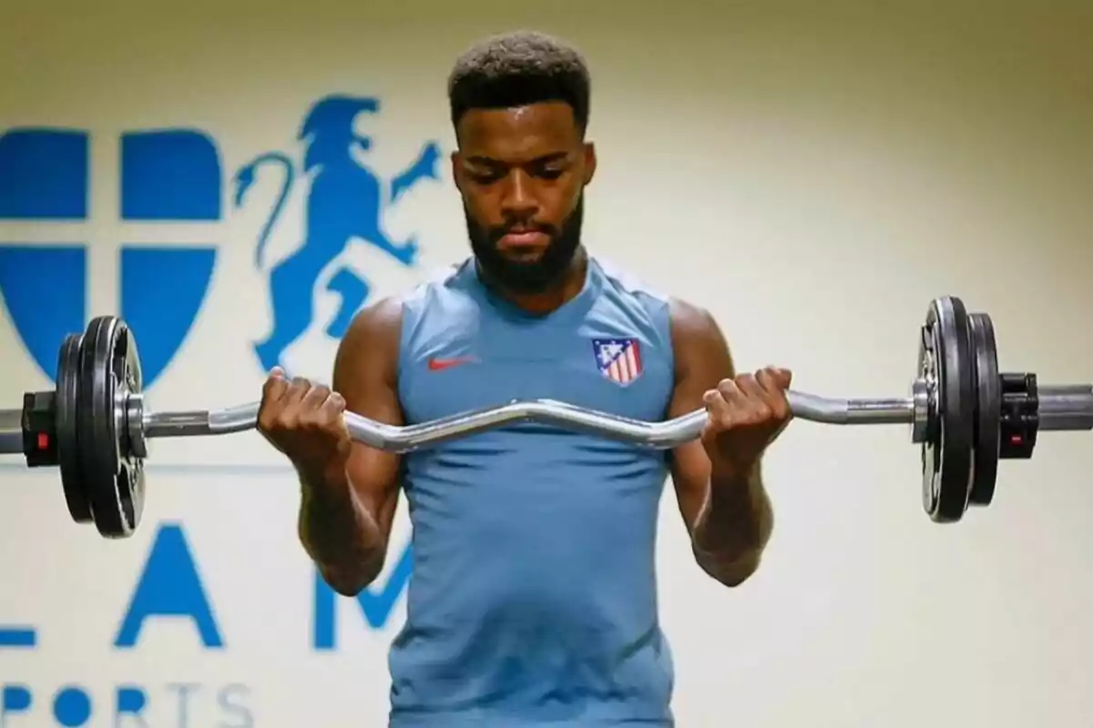 Un hombre levantando pesas con una barra en un gimnasio, usando una camiseta sin mangas del Atlético de Madrid.