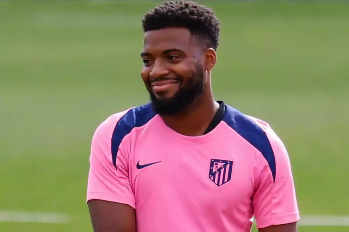 Un jugador de fútbol con camiseta rosa del Atlético de Madrid sonríe en un campo de entrenamiento.