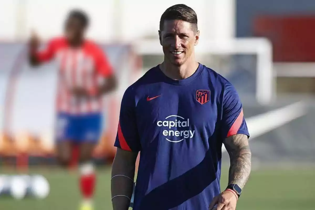 Un hombre con camiseta de entrenamiento del Atlético de Madrid sonríe mientras camina en un campo de fútbol.