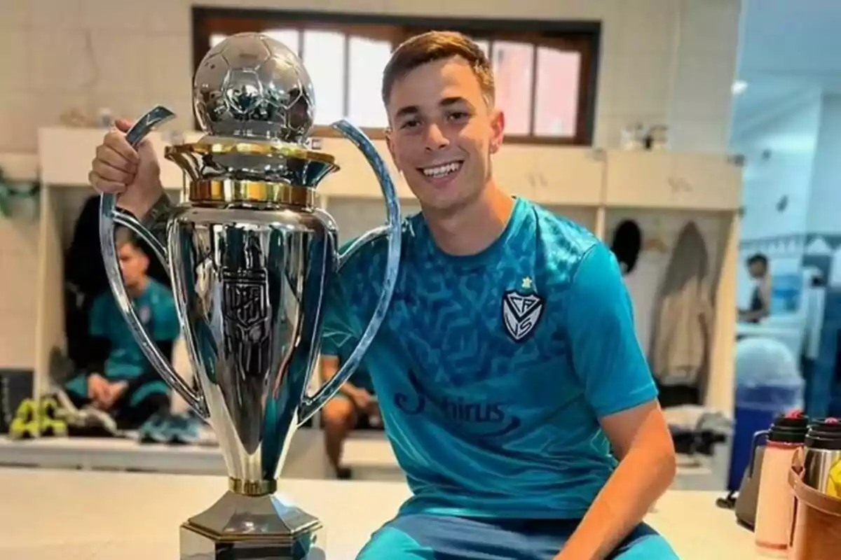 Un jugador de fútbol sonríe mientras sostiene un gran trofeo plateado en un vestuario.