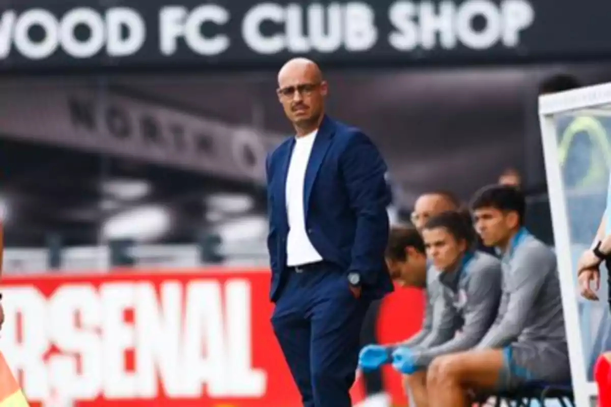 Un entrenador de fútbol de pie junto al campo, con jugadores sentados en el banquillo detrás de él y un cartel de "Arsenal" en el fondo.